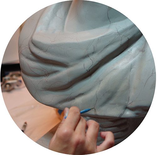 A hand with a fine brush painting thin veining lines onto the folds of a pale blue-gray sculpted piece.