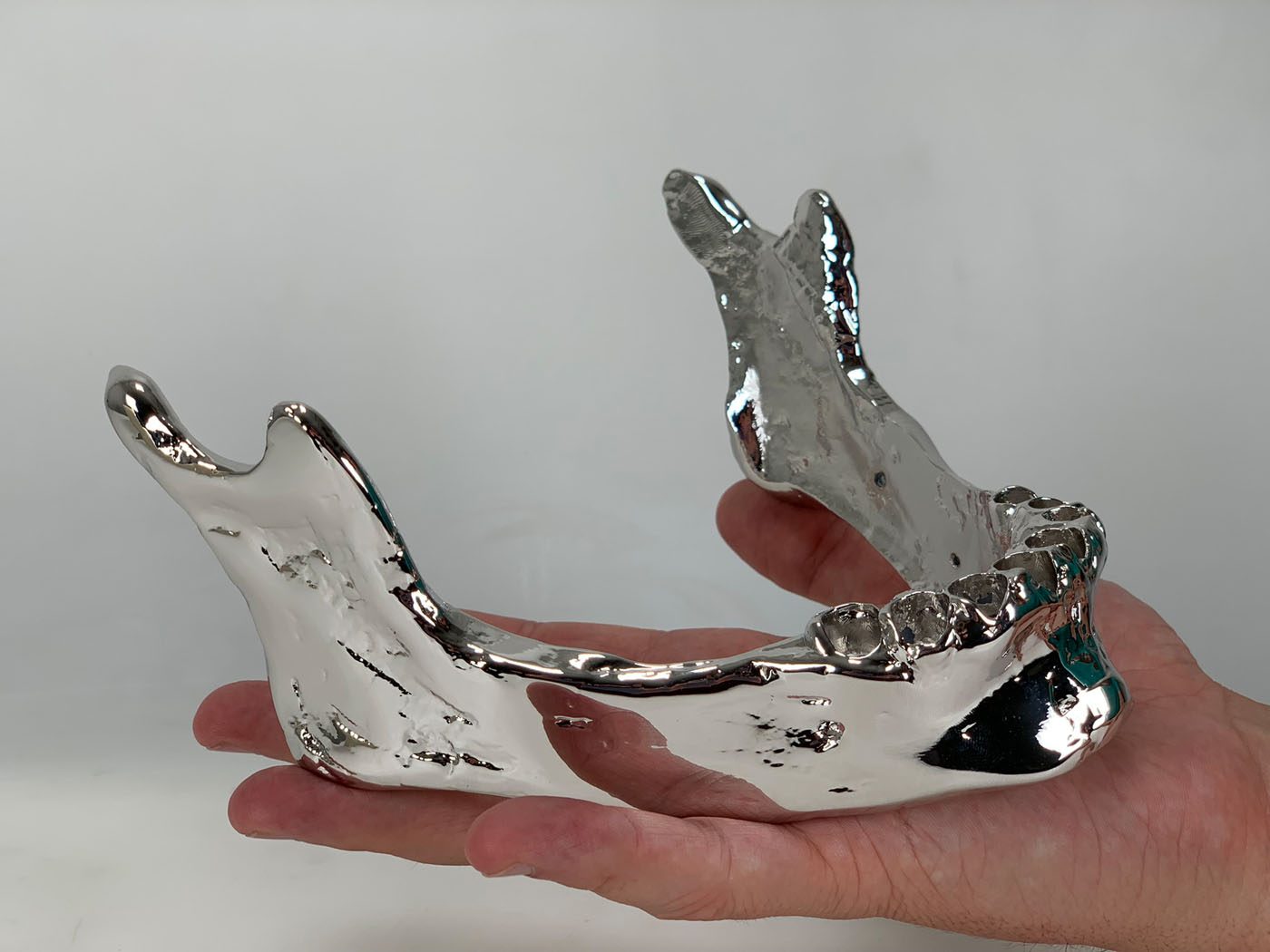 Mirror chrome plated jawbone held on an open palm against a light grey background, showing bright reflective surfaces