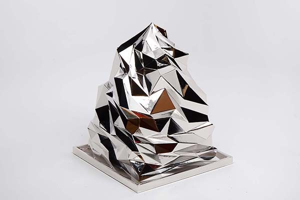 Faceted mirror-finish sculpture shaped like a small mountain sitting on a shallow square base against a white backdrop