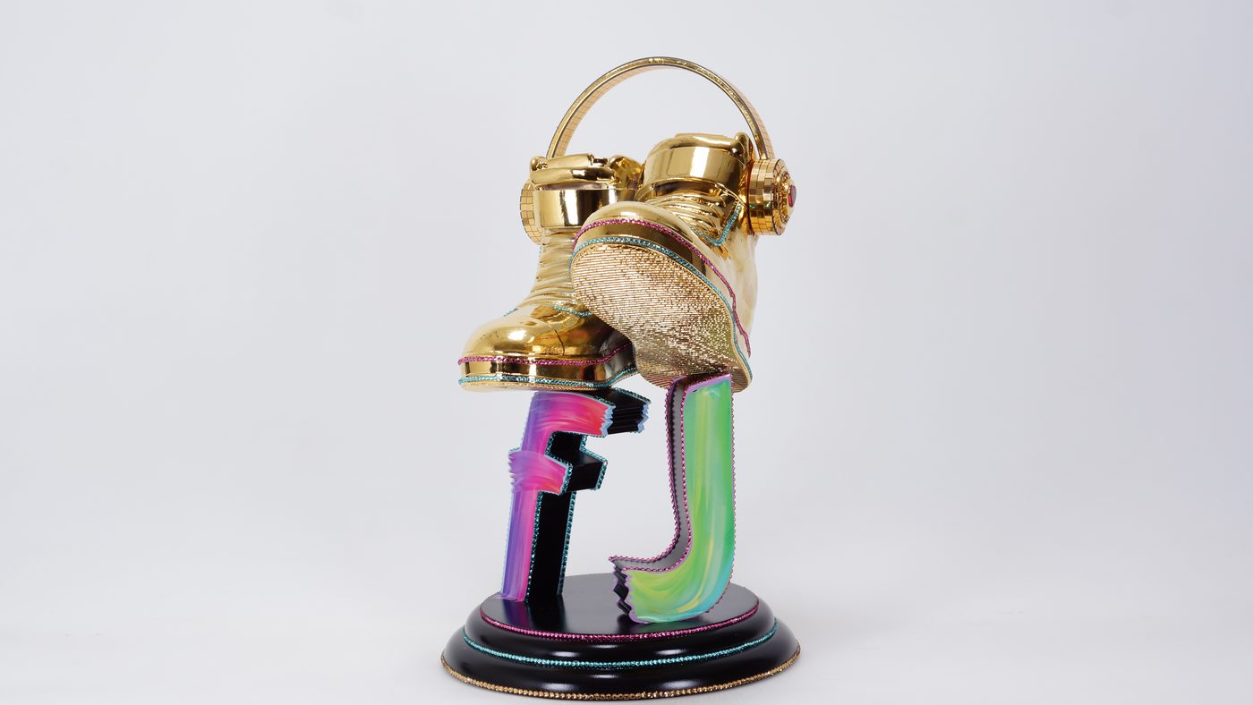 Glossy gold sneakers wearing headphones, mounted above colorful letter shapes on a black round base with rhinestone trim