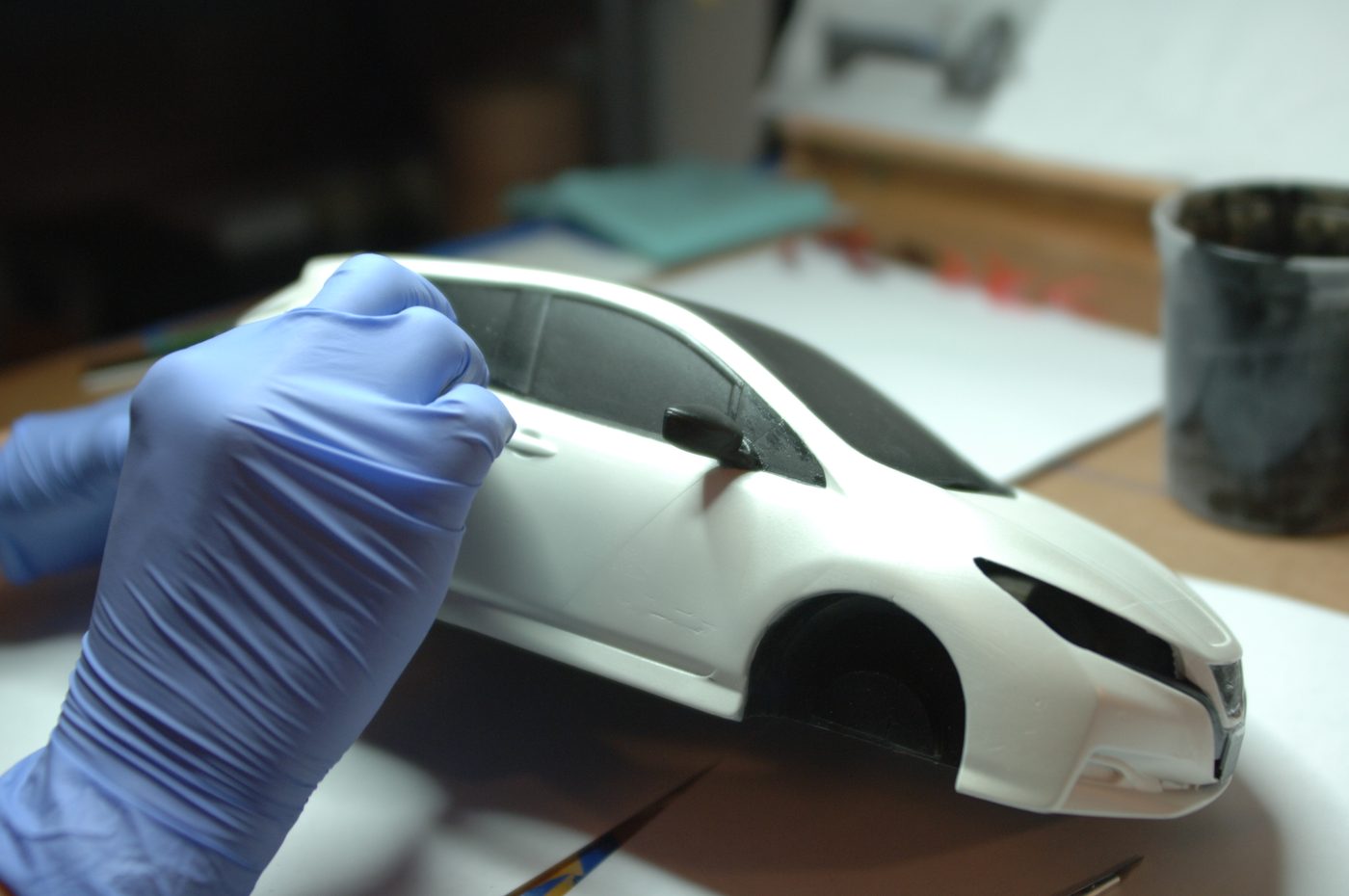 Gloved hand detail-painting the black window trim on a white scale car model with a fine brush at a workbench