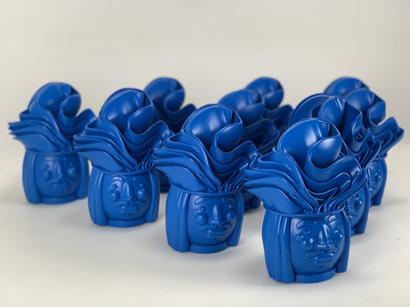Group of ten matching blue-painted character head sculptures with sculpted hair arranged in rows on a white surface