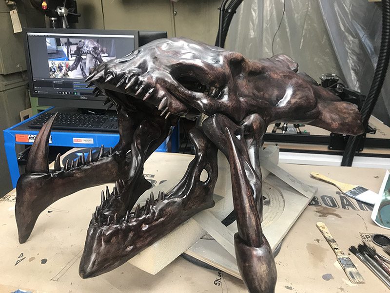 Large dark metallic-painted creature skull sculpture sitting on a workshop table with paint brushes and a computer behind it