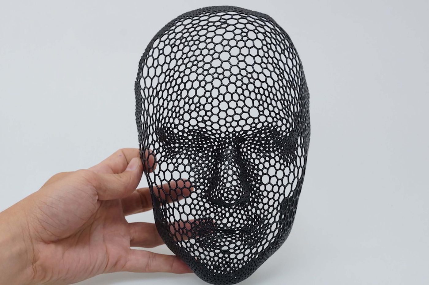 Hand holding a black 3D printed mask made of a fine honeycomb lattice, viewed from the front against a plain light background