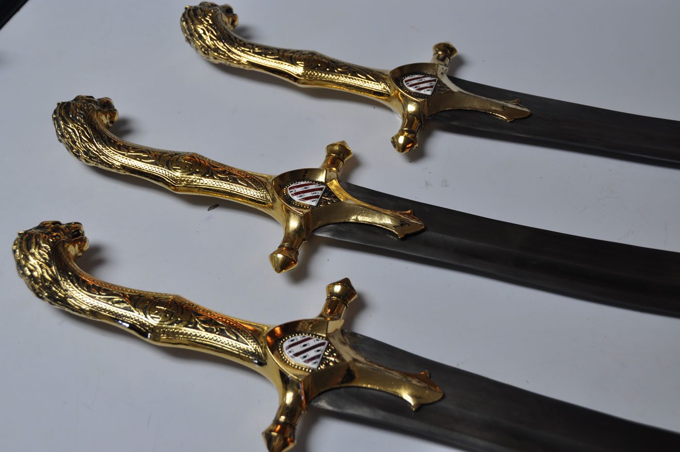 Three ornate gold-finished sword hilts with lion-head pommels and heraldic shields, blades lying on a light surface