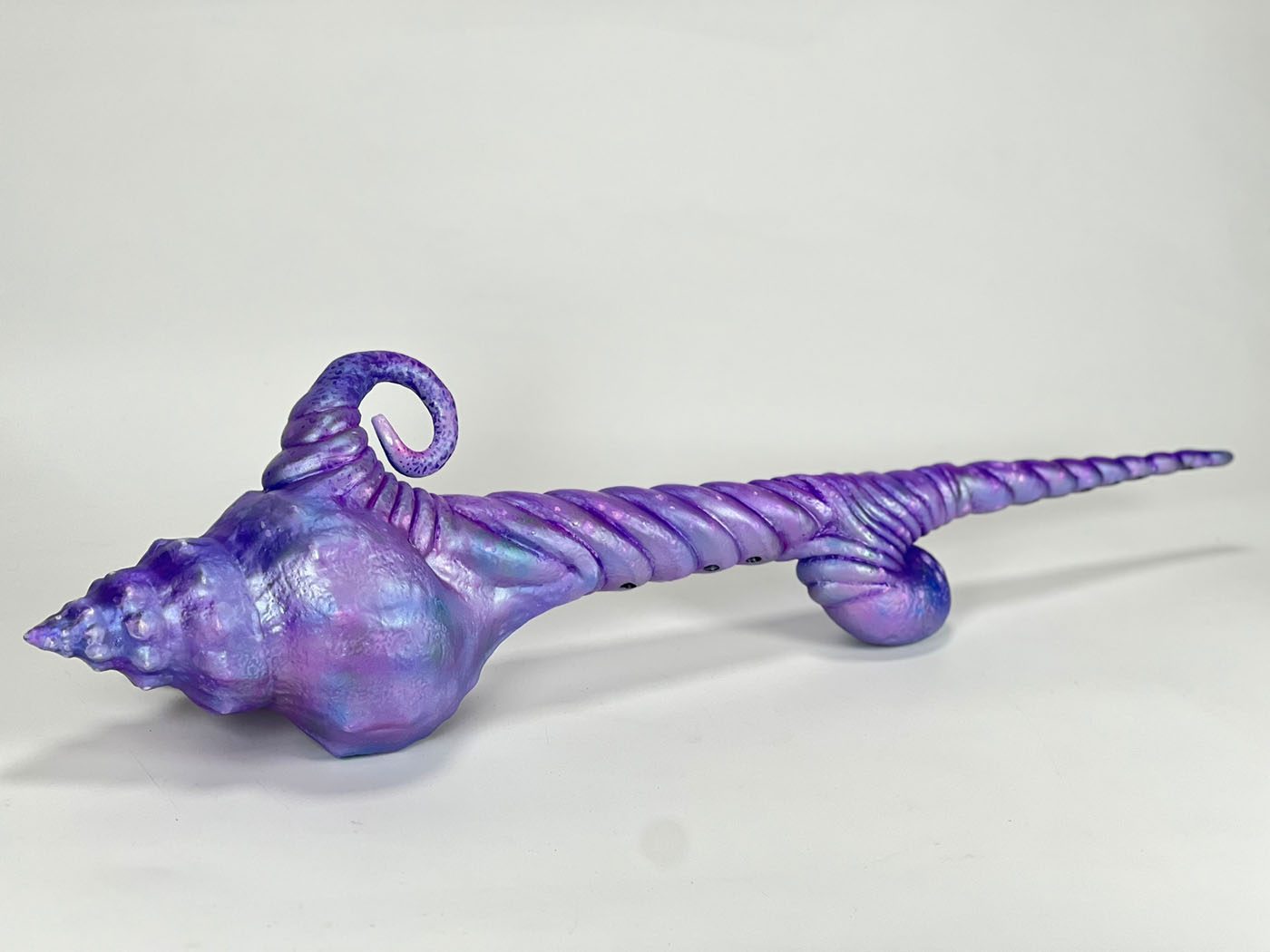 Purple and blue iridescent conch-shell horn prop with a spiral tail and curled hook, resting on a light gray surface