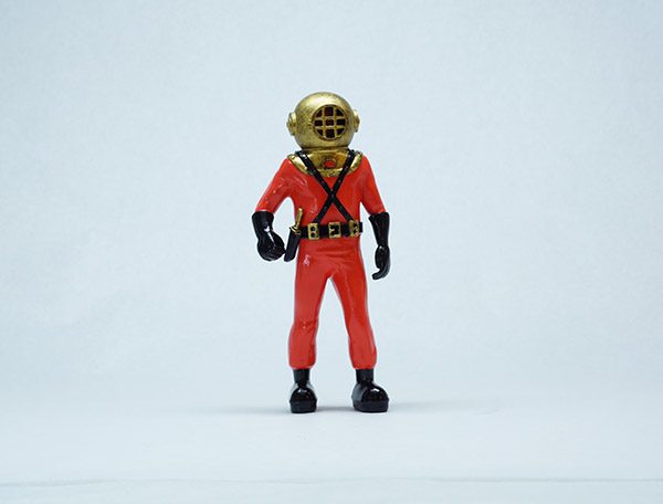 Small red-suited diver figure with a gold helmet and black gloves and boots, standing against a pale blue-gray backdrop
