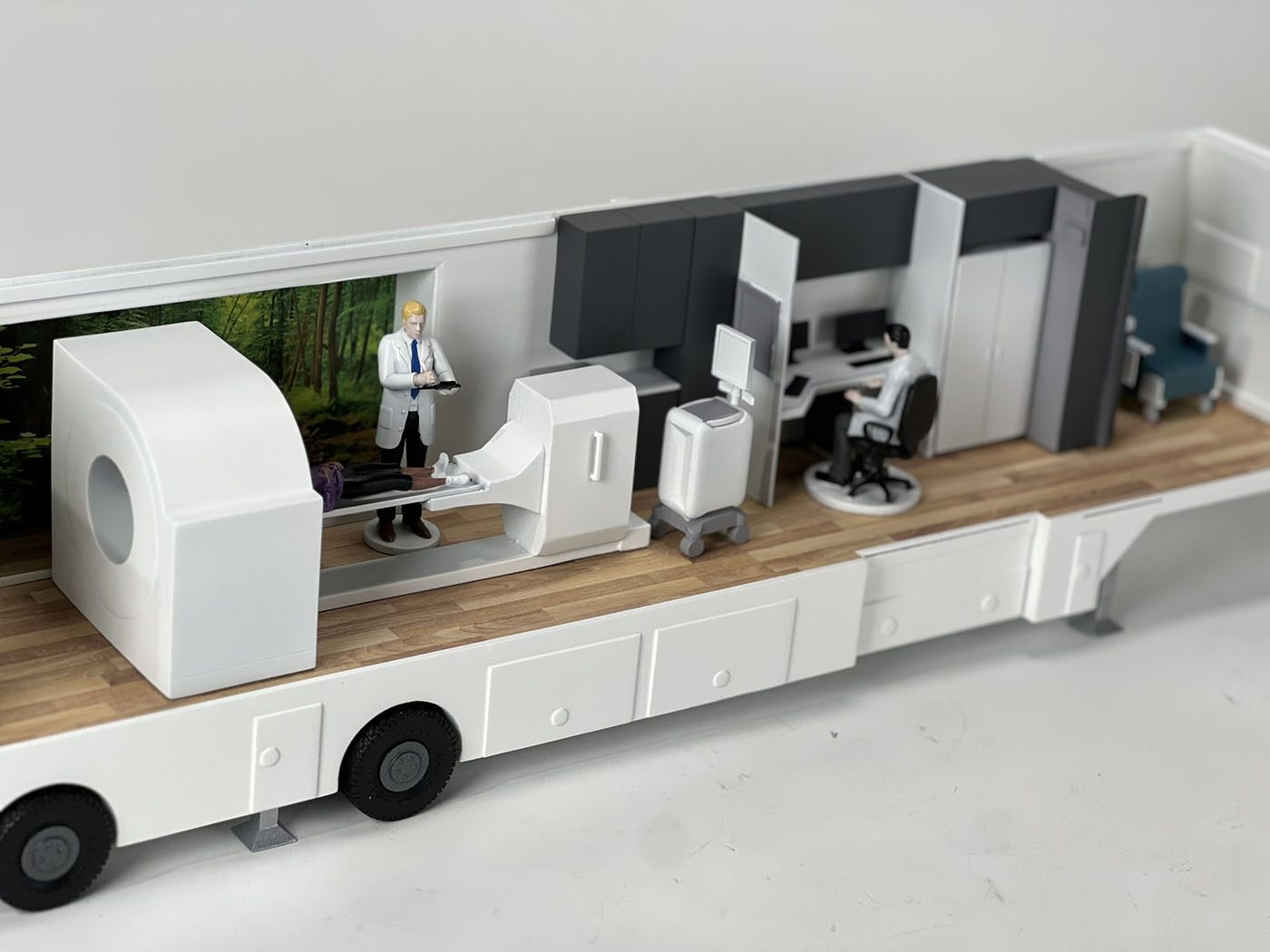Cutaway scale model of a mobile medical trailer showing an imaging machine, control station and wood-look floor inside a white shell