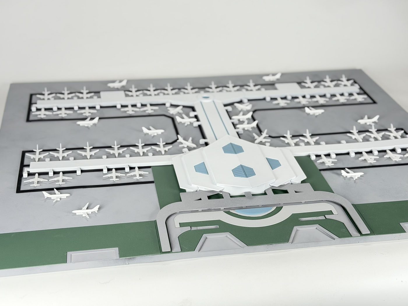White airport terminal scale model with concourses and small aircraft on a gray and green painted site base