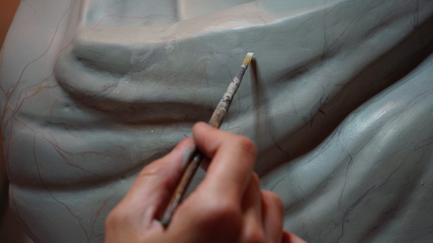 Hand holding a fine paintbrush over a large gray sculpture, painting thin marble-style veining along the folds
