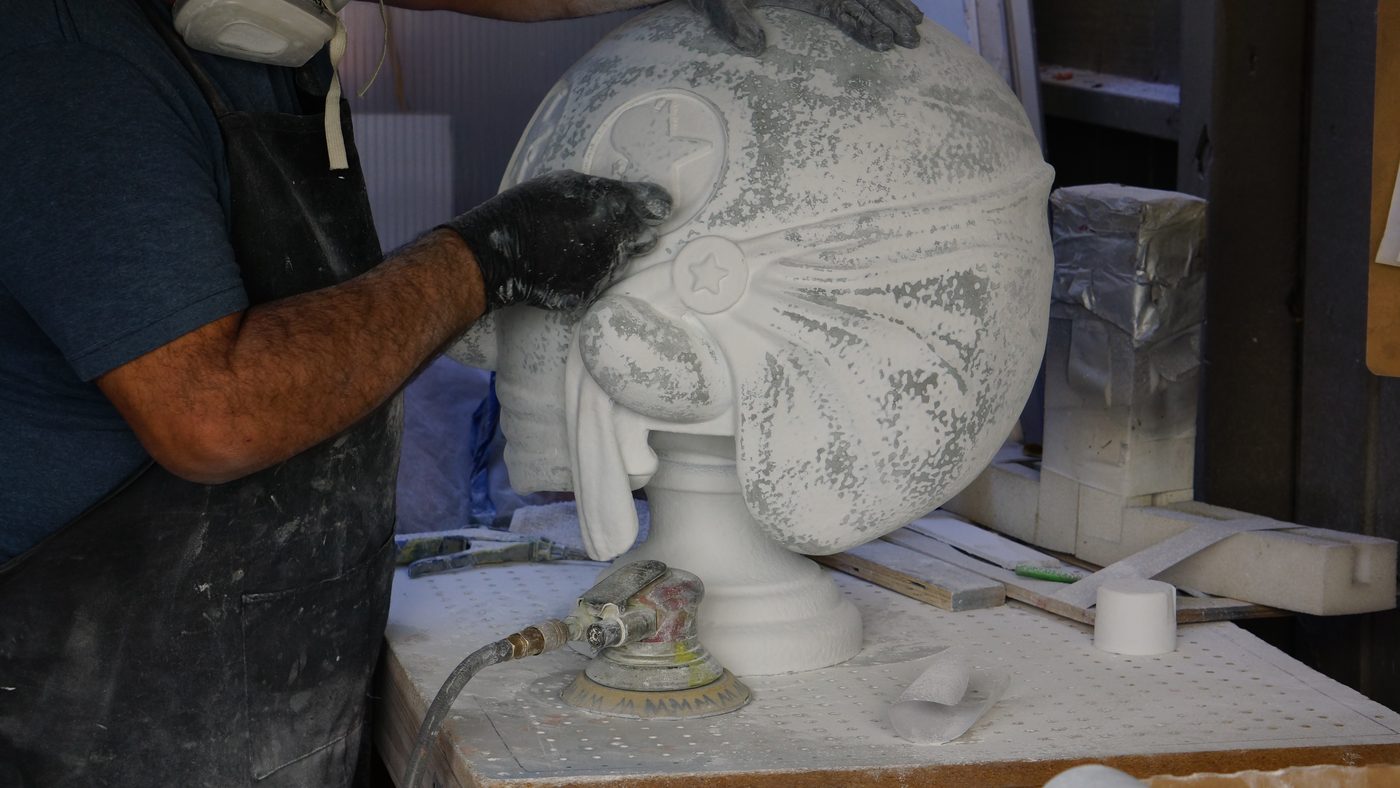 Worker in a respirator and black gloves sanding a large white sculpture on a workbench with a pneumatic sander beside it