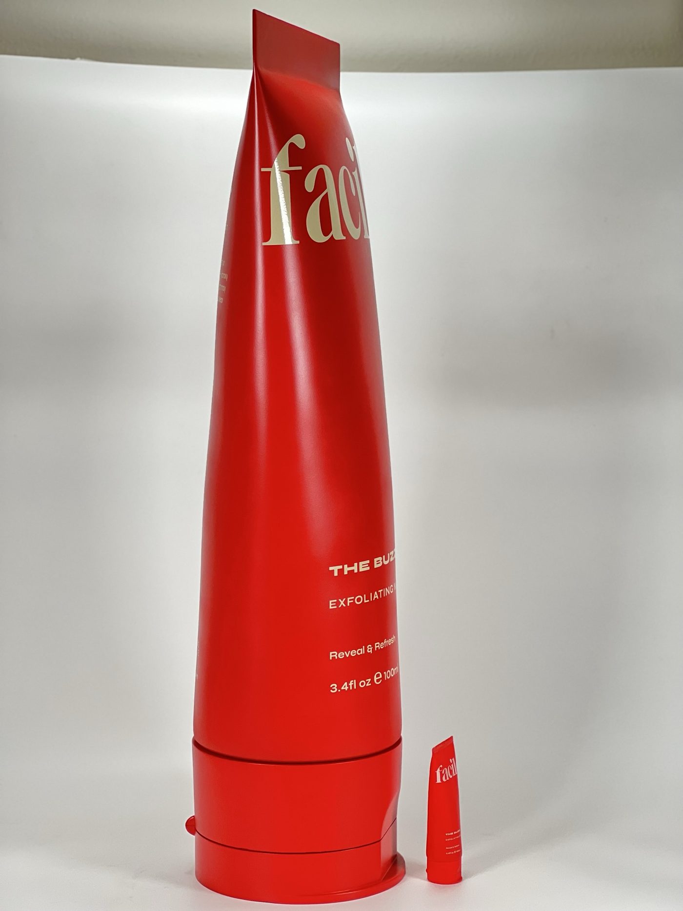 Tall matte red squeeze-tube replica with cream lettering standing beside a tiny actual-size tube on a white surface