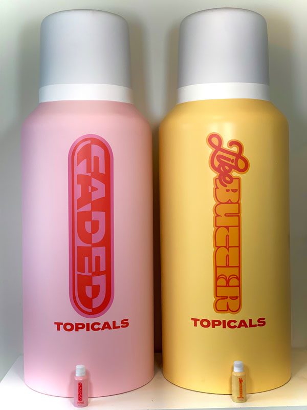 Two large pink and yellow spray-can replicas with gray caps and bold graphics, each with a small actual-size bottle at its base
