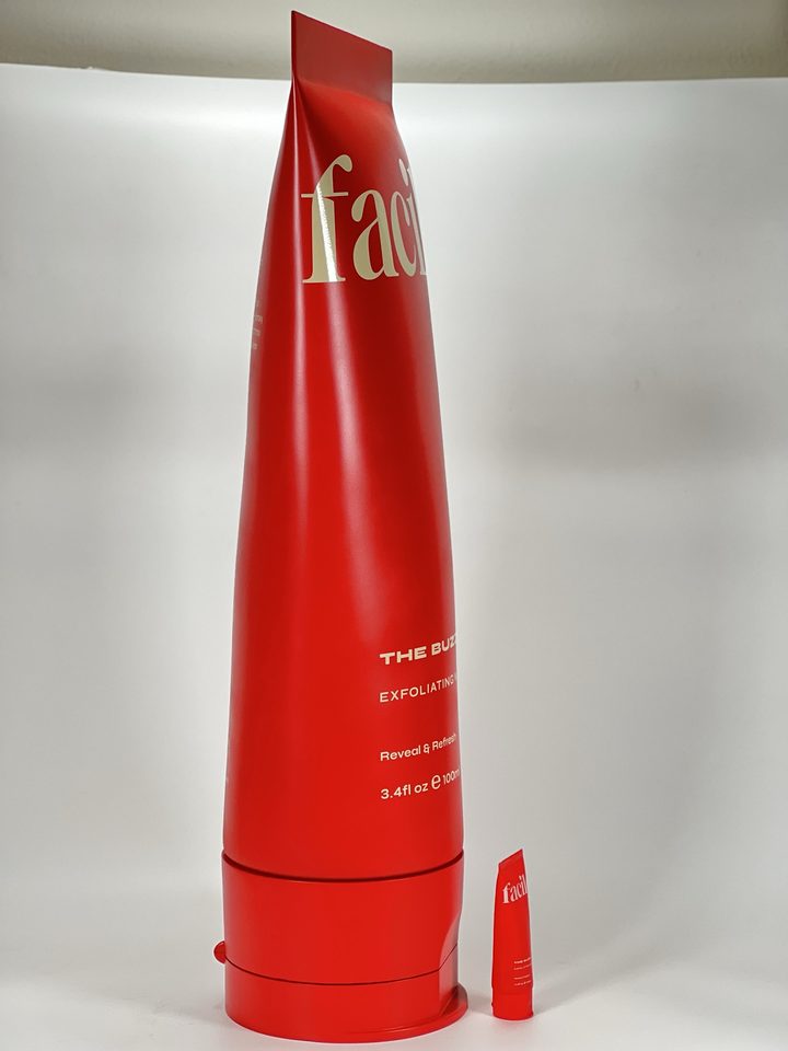 Tall matte red squeeze-tube replica with cream lettering standing beside a tiny actual-size tube on a white surface