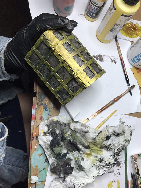 Gloved hand holding a small gold and green painted treasure chest prop over a table of brushes, paint bottles and a stained cloth