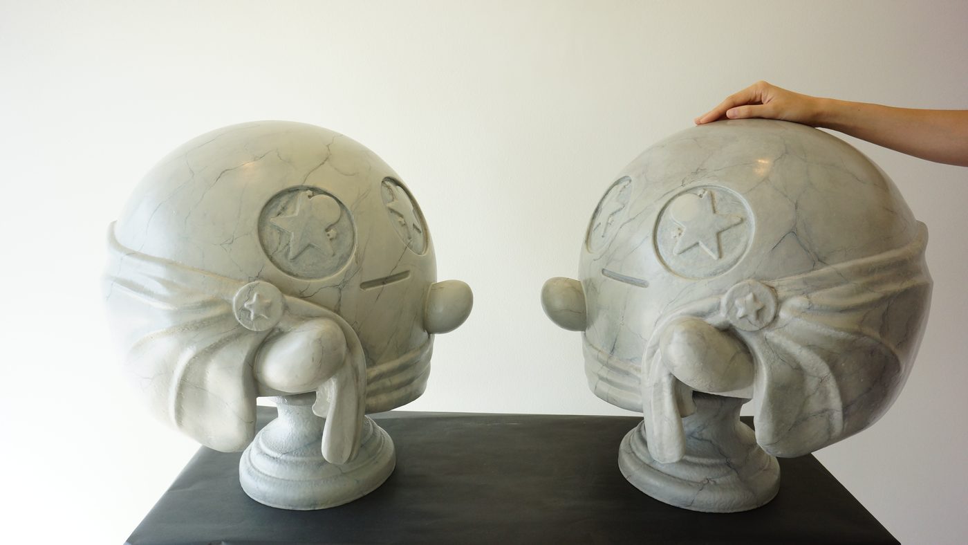 Two large sculpted helmets with star emblems painted to look like grey veined marble, with a hand resting on one for scale