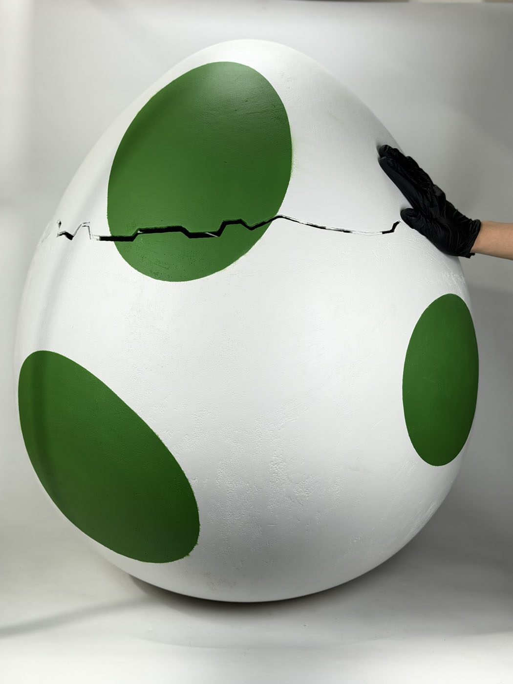 Oversized white egg prop with large green spots and a jagged crack line, a gloved hand resting on its side for scale