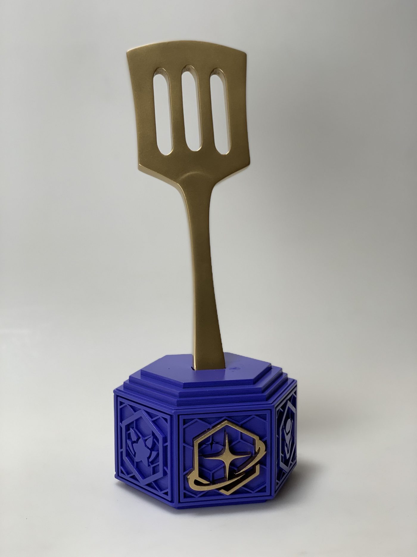 Gold spatula-shaped trophy standing in a hexagonal purple base with raised decorative panels and a gold emblem