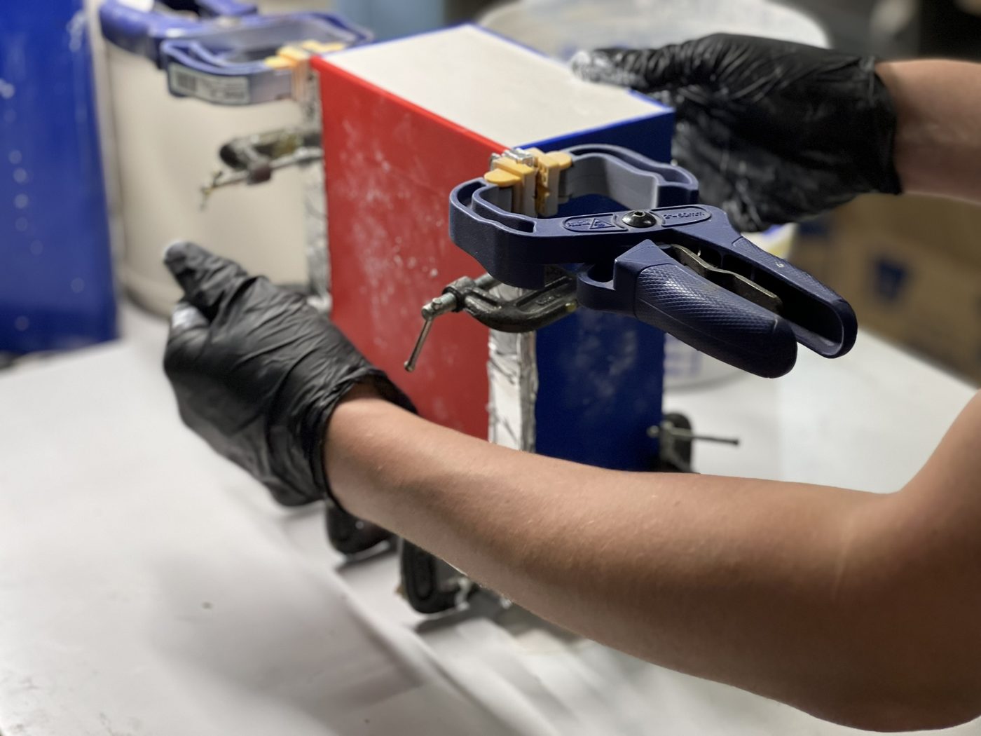 Gloved hands holding a clamped red and white mold box on a workbench while it is assembled for a silicone pour.