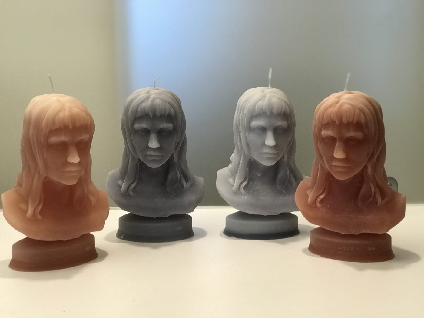 Four bust-shaped candles of a long-haired figure in two colors, peach and gray, lined up on a white surface.