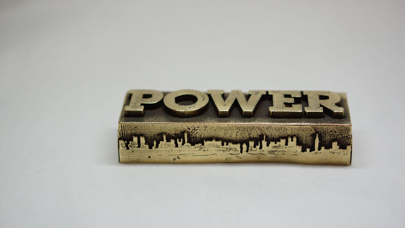 Brass-toned block with raised serif lettering on top and a city skyline cut along its edge, on a plain light gray background