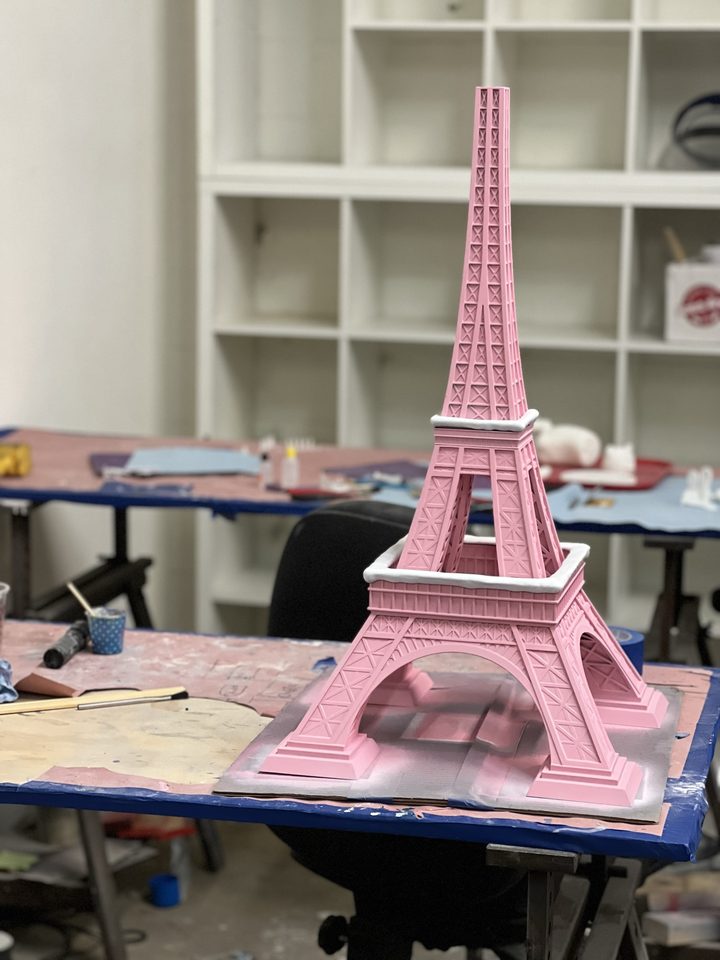 Pink painted Eiffel Tower model on a paint-stained workshop bench with white shelving behind it